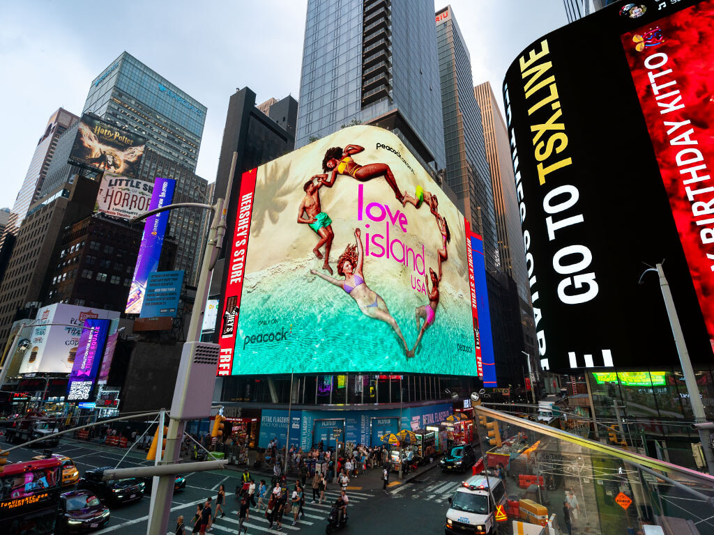 #94. Biển quảng cáo khổng lồ giữa khu phố đông đúc hiển thị hình ảnh bãi biển với những người tạo thành hình trái tim cùng dòng chữ “love island USA” nổi bật.
